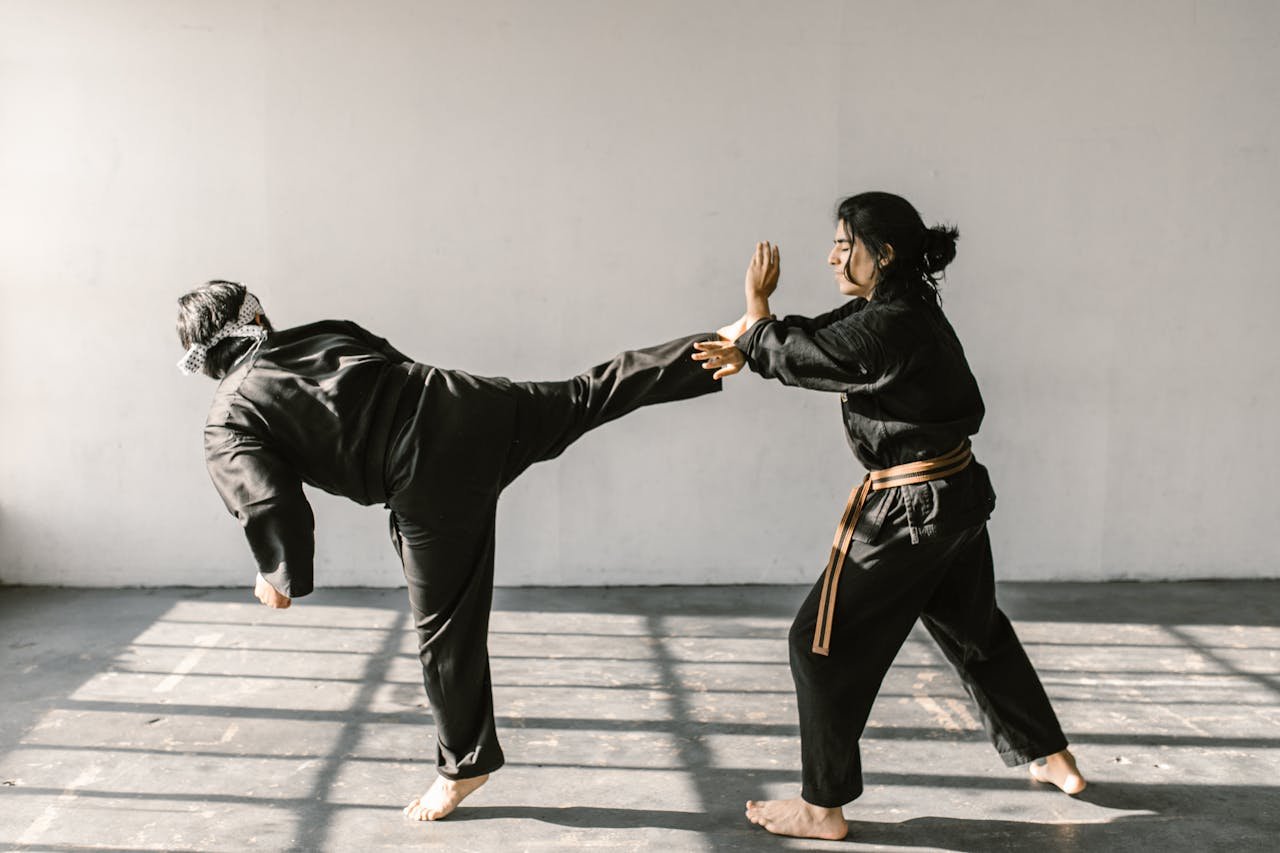 Two martial artists practicing karate kicks and self-defense in a sunlit room.